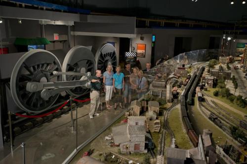 A group of people stands in front of a large model train display with gears and buildings in a museum setting.