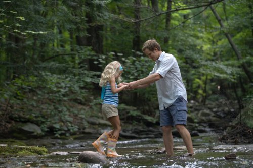 A man helps a young girl balance while playing in a shallow stream surrounded by lush green trees.