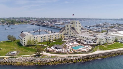 Aerial view of a waterfront hotel with a pool, marina, and boats in the background on a sunny day.