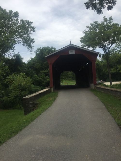 A red covered bridge spans a path surrounded by greenery under a cloudy sky.