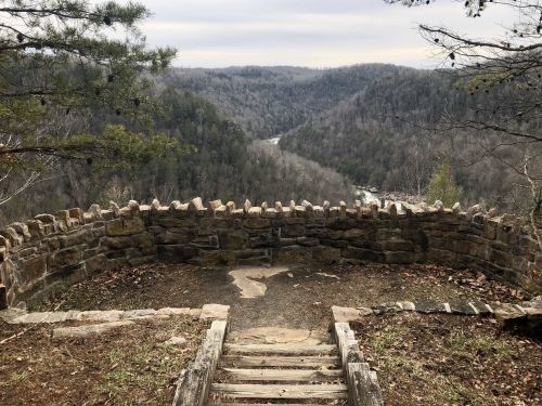 Scenic overlook with a stone wall, featuring a view of a valley and river surrounded by wooded hills.