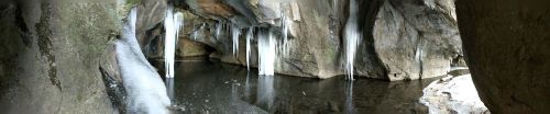 A panoramic view of a cave with icicles hanging from the rocky ceiling above a dark, still pool of water.