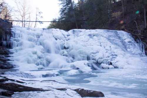 Frozen waterfall with icy cascades and a serene winter landscape, surrounded by trees and a distant bridge.