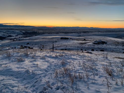 A snowy landscape at sunset, with rolling hills and distant mountains under a colorful sky.