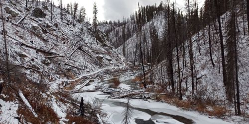 A snowy landscape with a frozen river, surrounded by barren trees and rocky hills.