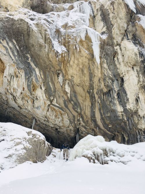 A person stands near a snow-covered cave entrance, surrounded by icy rock formations and icicles hanging above.