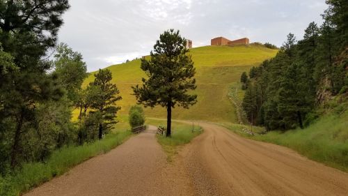 A dirt road splits into two paths, surrounded by green hills and trees under a cloudy sky.