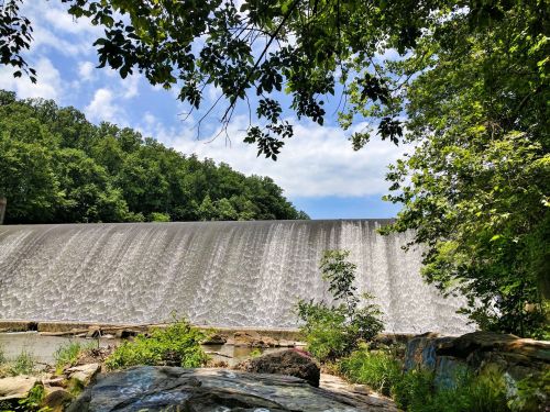 A serene view of a waterfall cascading over a dam, surrounded by lush greenery and a blue sky.