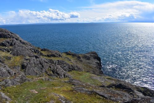 A rocky coastline meets a calm blue sea under a bright sky with scattered clouds.