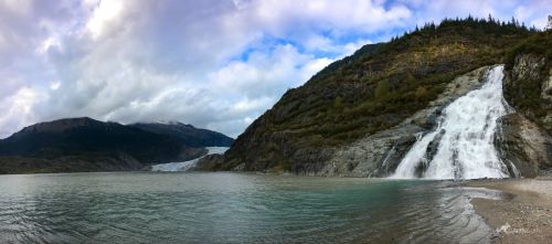 A panoramic view of a waterfall cascading into a serene lake, surrounded by mountains and lush greenery under a cloudy sky.