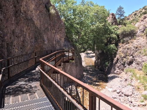 A winding metal walkway alongside a rocky canyon and a shallow stream, surrounded by greenery and cliffs.