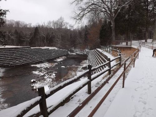 Snow-covered landscape with a stream, stone steps, and a wooden railing in a wooded area.