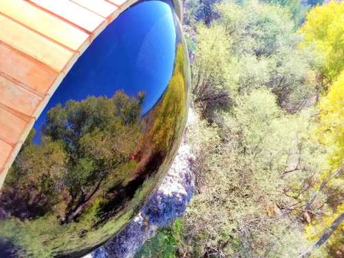 A reflective black sphere perched on a rocky ledge, surrounded by lush green trees and a clear blue sky.