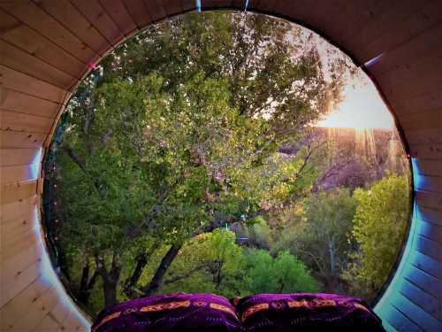 View from a circular window showcasing lush trees and a sunset, with cozy bedding in the foreground.