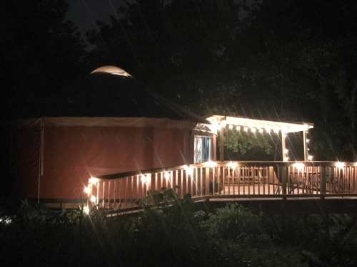 A cozy yurt illuminated by string lights at night, surrounded by greenery.