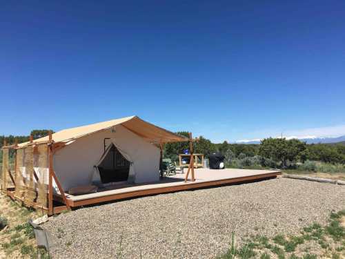 A spacious glamping tent on a wooden deck, surrounded by greenery and mountains under a clear blue sky.