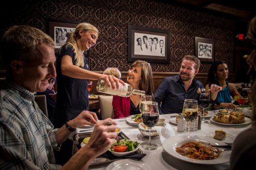 A server pours wine for guests at a lively dinner table filled with food and drinks in an elegant restaurant setting.