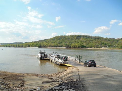 A ferry docked at a riverbank with a car waiting to board, surrounded by green hills and a blue sky.
