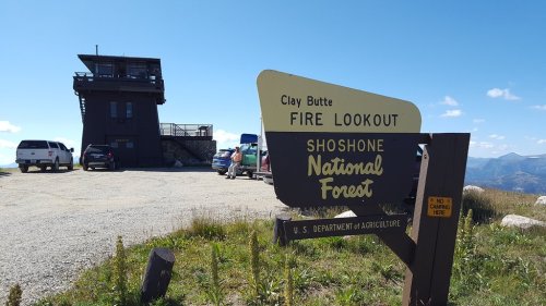 Sign for Clay Butte Fire Lookout in Shoshone National Forest, with a lookout tower and parked cars in the background.