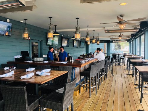 A coastal restaurant interior with wooden tables, bar area, and two staff members chatting. Large windows overlook the view.