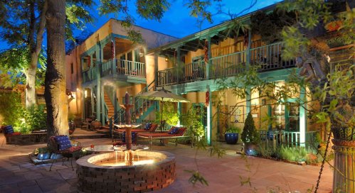 A cozy courtyard with a fire pit, surrounded by colorful seating and lush greenery, illuminated at dusk.