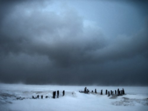 A desolate winter landscape with dark clouds and weathered wooden posts emerging from a snowy ground.