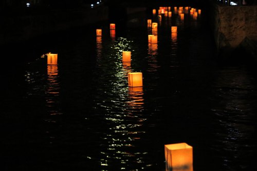 Glowing lanterns float on a dark water surface, reflecting light in a serene nighttime scene.
