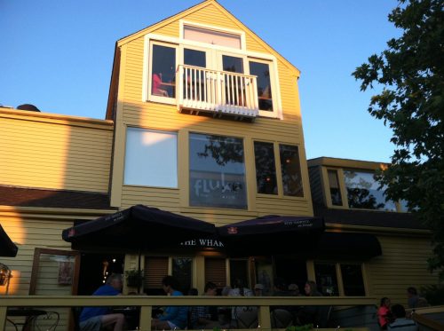 A yellow building with large windows and a balcony, featuring outdoor seating and umbrellas at a restaurant.