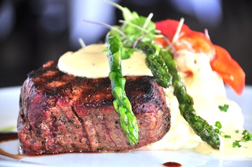 A perfectly cooked steak topped with asparagus and a creamy sauce, served with mashed potatoes and a slice of tomato.