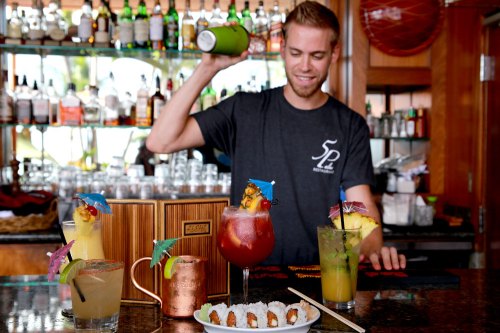 A bartender shakes a cocktail behind a bar with colorful drinks and sushi on the counter.