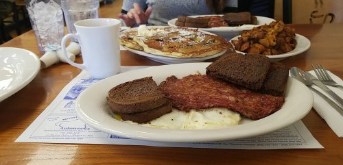 A plate with corned beef hash, eggs, and rye bread, with pancakes and home fries in the background. Coffee cup on the side.
