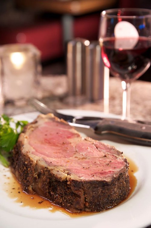 A succulent slice of prime rib on a plate, garnished with greens, accompanied by a glass of red wine.