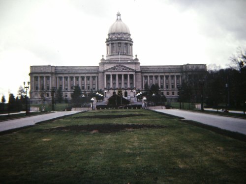 A large, historic government building with a dome, surrounded by landscaped grounds and a cloudy sky.