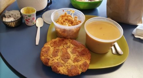 A green plate with a large cornbread muffin, macaroni and cheese, and a bowl of soup on a blue table.
