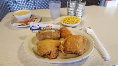 A plate with fried chicken, a baked potato, corn, and a side of sour cream, with soup and water in the background.