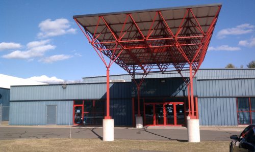 Modern building entrance with a large red canopy and blue exterior, set against a clear blue sky.