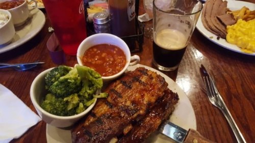 A plate of grilled ribs with barbecue sauce, broccoli, baked beans, and a glass of dark beer on a wooden table.