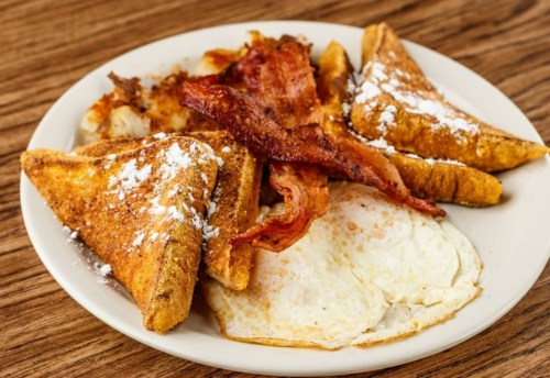 A plate of breakfast featuring French toast, crispy bacon, and fried eggs, garnished with powdered sugar.