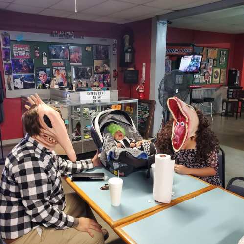 Three people wearing oversized, whimsical masks sit at a table, with a baby in a car seat between them.