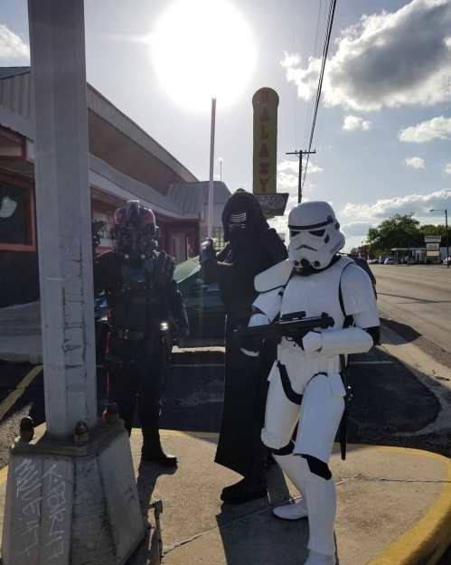 Three costumed characters, including a stormtrooper and a figure in black, pose under a bright sun near a diner.