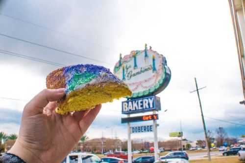A hand holds a colorful king cake in front of a bakery sign, with cloudy skies and cars in the background.