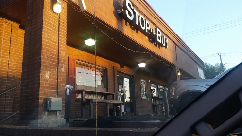 Exterior of a store named "Stop and Buy," featuring brick walls and outdoor seating, with warm evening lighting.