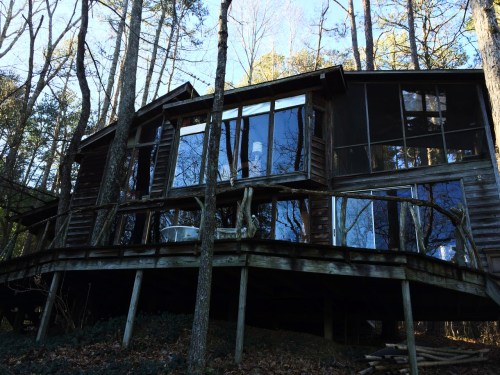 A modern wooden cabin with large windows, nestled among trees, featuring a raised deck and a serene forest backdrop.