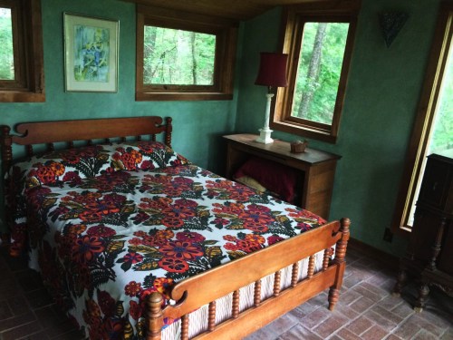Cozy bedroom with a floral bedspread, wooden furniture, and large windows overlooking a green forest.