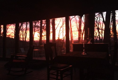 A cozy interior with a table and rocking chair, framed by large windows showcasing a vibrant sunset through trees.