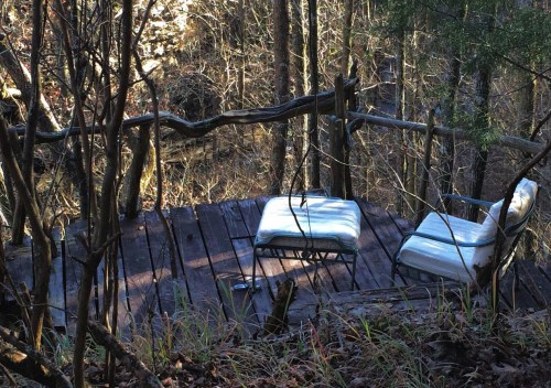 A wooden deck in a forest, featuring two empty chairs surrounded by trees and foliage.