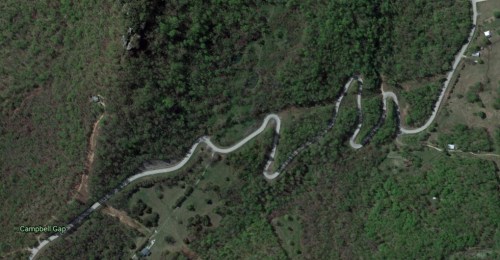 Aerial view of a winding road through lush green hills, with a few buildings visible nearby.