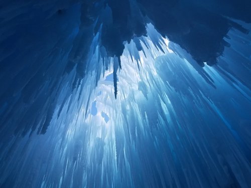 Icicles hang from a ceiling of ice, illuminated by soft blue light, creating a stunning, ethereal atmosphere.