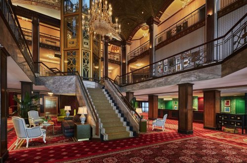 Elegant hotel lobby featuring a grand staircase, chandelier, and plush seating in a richly decorated space.