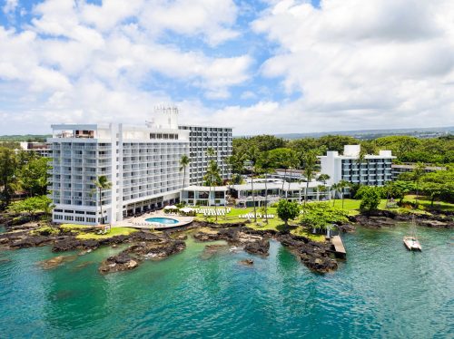 Aerial view of a modern hotel by the water, surrounded by greenery and rocky shoreline under a partly cloudy sky.
